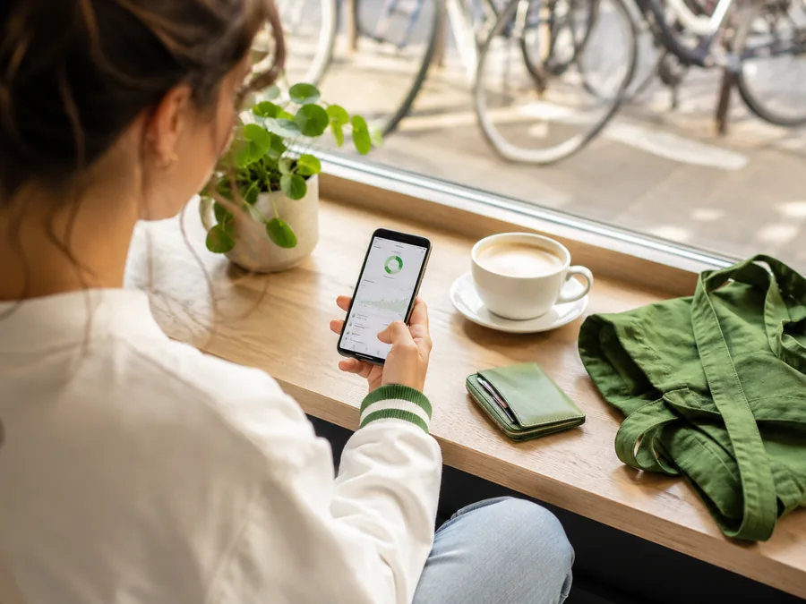 Frau prüft ihre Finanz-App am Smartphone, daneben Kaffee und Portemonnaie